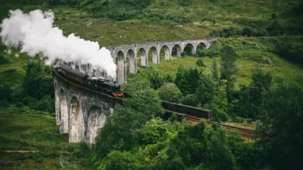 A dramatic shot of the vintage Jacobite steam train crossing the iconic, curved Glenfinnan Viaduct in the rugged and misty Scottish Highlands, with Loch Shiel in the background.